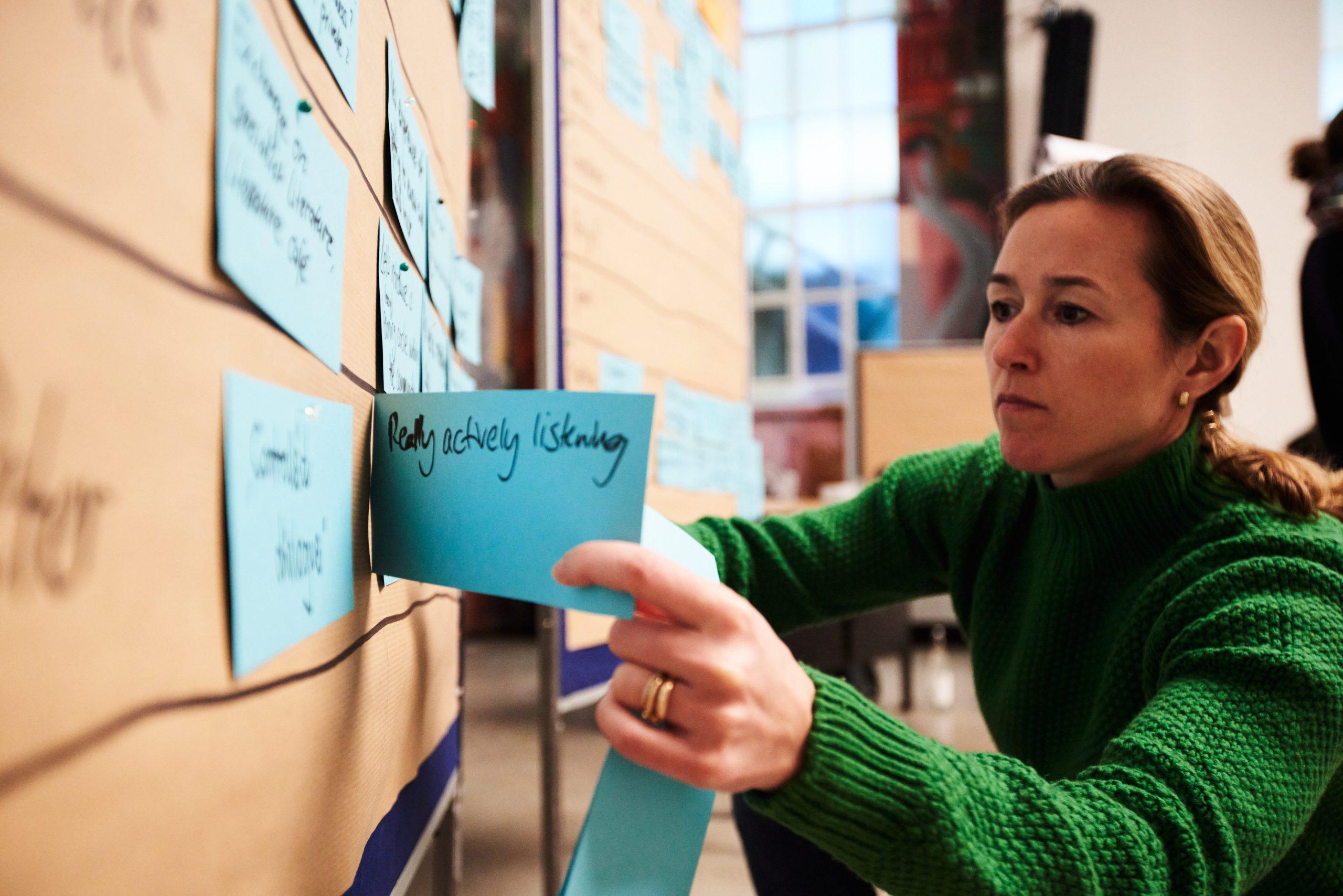 Person in grünem Pullover klebt blaue Notizzettel mit handschriftlichen Texten an eine Wand